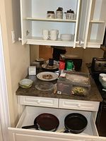 Kitchen countertop and open shelves showing mugs, cups, plates, wood bread slicer, Pyrex dish, and partially visible Sony radio.