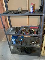 Full view of grey metal shelving unit with multiple shelves holding assorted hand tools including pliers, snips, planes, electric drill, stone, hammers, screwdrivers, chisels, sockets, stapler, paint and more.
