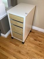 Front angle view showing four wood-toned drawers in a white cabinet on caster wheels.