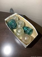 Box containing six glass insulators in clear and greenish-blue colors with various shapes and sizes.