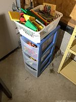 Blue 3-drawer plastic organizer with white basket on top filled with Lego and toy parts, in basement setting.