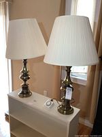 Pair of metal table lamps standing on a white shelf in a room with beige walls and window.