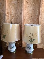 Pair of lamps with Dornbush style cream-colored lampshades with floral and fern designs, placed on wooden surface against an OSB wall