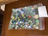 Clear plastic bag filled with a diverse collection of small and large glass marbles featuring various colorful designs including swirls and solid colors.