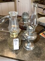 Three oil lamps displayed on kitchen counter; two glass and one metal. One glass lamp is Aladdin style without chimney, tallest lamp measures 17 inches.