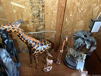 Collection of safari animals on wooden surface against wall, showing large leather giraffe in foreground, smaller wood giraffes, wood antelope, and resin zebra candle holder with small plant on base.