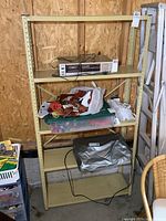 Front view of beige metal shelf unit showing four shelves with various items placed on them, including electronic device, toys, plastic bins, and bags. Shelf is positioned against a wooden wall.