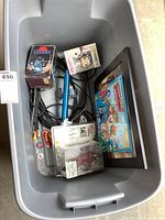 View of the lot contents in a plastic tote, showing Star Wars Trilogy box, Funko Pop figure, framed Justice League calendar image, wizard-style wand, various cables, watch, and other small items.