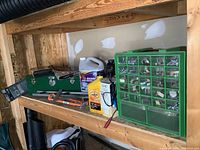 View of wooden shelving with a green plastic hardware organizer filled with assorted hardware items, alongside automotive fluids, tools, and a green metal toolbox