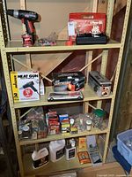 Yellow metal shelving unit holding various tools and hardware items including cordless drill, heat gun, air compressor, soldering kit, paint stripper, and assorted small containers and boxes.