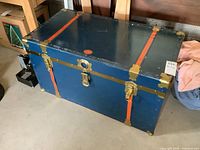 Full front view of closed vintage metal trunk showing blue metal body, orange straps, brass latches, and keyhole.