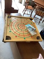 Top view of vintage wooden carrom board with concentric red and green circles and four corner pockets with netting; plastic bag of playing pieces on board