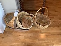 Multiple wicker baskets placed on a wooden floor and in a dark brown woven tray. Shows different shapes and sizes including oval and round baskets with handles.