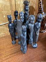 Eight dark wood carved statues of 10 to 15 inch height with individual features on a wooden surface against particle board background.