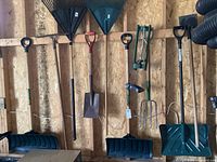 Wide shot of various yard tools hanging on plywood wall showing snow shovels, leaf rakes, metal rake, fork, shovel, and sprinkler