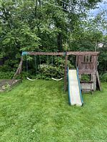 Full front view of the wooden playhouse with two swings and slide on a grassy lawn surrounded by trees.