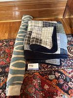 Rolled-up rug with modern blue and beige pattern beside folded patchwork comforter on a patterned red carpet.