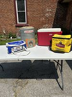 All 4 coolers on a white folding table outdoors: blue Labatt's soft cooler, cylindrical Arctic metal cooler, red/white Coleman plastic cooler, yellow Sleeman soft cooler.