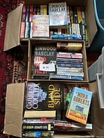 Three boxes of mystery fiction novels stacked with visible titles by Linwood Barclay, Nora Roberts, Catherine Coulter, and Julie Garwood.