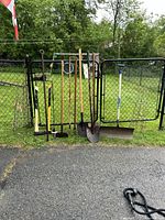 Photo showing multiple garden tools leaning against a chain link fence, including axe, sledgehammer, shovels, rakes, hoes, and a metal wire plant support.