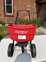 Front view of EarthWay EV-N-SPRED red hopper seed spreader with two large black wheels and black frame on white table outside.