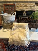 Photo of round woven wicker basket, rectangular dark brown woven wicker basket, and folded white Indigo Home throw blanket on table with stone wall background.