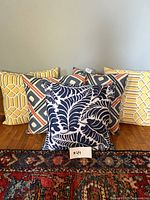 Five cushions arranged in two rows, three cushions in the back row with geometric patterns in yellow and multicolor, two cushions in front with navy blue botanical leaf pattern and yellow geometric pattern. Cushions show some marks and signs of wear.
