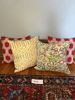 Four indoor/outdoor cushions arranged side by side, showing front views including two red hexagonal pattern cushions, yellow geometric pattern cushion with marks, and multicolored floral cushion.