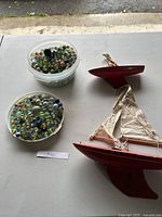 Two containers of assorted marbles next to two vintage red wooden toy sailboats on a white table.