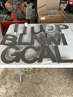 Photo showing multiple metal letters arranged on a table in a garage environment. Letters visible include I, U, D, P, B, L, N, M, G, C, A.