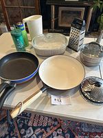 All items in the kitchen lot displayed on table including pans, CorningWare dish, cheese grater, steamers, milk pitcher, and cups.