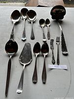 Top-down view of 12 miscellaneous silverware serving pieces arranged on a surface showing various types of spoons, a serving fork, and a pie server.