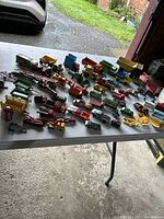 Wide angle photo showing about 45 toy vehicles on a white table in a garage including cars, trucks, and construction vehicles of various brands and colors.
