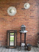 Wide view showing two tin sunflowers mounted on brick wall, mirror leaning against wall, tall wooden table with lantern on top and copper pot on floor.