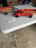 Two vintage metal toy trucks with farm wagon beds, one red and one grey, positioned side by side on a white table.