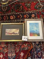 Two framed paper artworks on a red patterned rug: left is a warm-tone abstract scene by Wendy Cain in a gold frame; right is an abstract colorful sky-like wash in a gray wood frame.