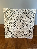 Full front view of square white carved wood wall art leaning against wall on hardwood floor.