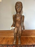 Frontal view of the tall wooden standing human sculpture on a hardwood floor against a white wall.