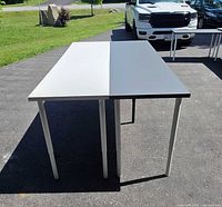 Two large fold-out work tables placed outside on pavement. One table has a grey top and the other a white top. Tables have thin metal legs adjustable for height.