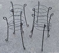 Four black metal plant stands standing on pavement, each with three curled legs and round top frames