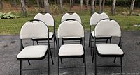 Front view of six black metal frame folding chairs with beige cushioned seats and backrests shown outdoors in an arranged row.