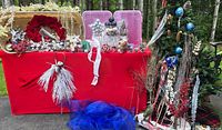 Wide view of holiday decoration collection showing picks, ribbons, ornaments, wreath, tree skirt, and decorations arranged on and in front of a red table cover outdoors.
