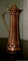 Front view of tall copper carafe with brass handle and lid showing embossed leaf motifs