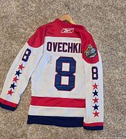 Overhead view of back of white Reebok NHL Winter Classic hockey jersey showing OVECHKIN name, number 8 in blue outlined in red, red shoulders and star patterns on sleeves, signature in black marker.