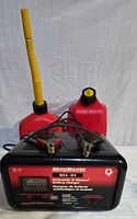 Front view of MotoMaster battery charger with two red gas cans behind it, showing yellow flexible spouts.