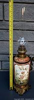 Full view of the oil lamp next to a measuring tape showing height of approximately 16 inches including glass chimney, ceramic hand-painted base and brass fittings.