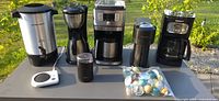 Full view of all machines and accessories on outdoor surface showing coffee urn, drip coffee makers, grinder, coffee pods, and warmer plate.