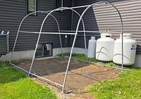 Full assembled metal tubular frame with arched supports placed on a grass lawn near a house with propane tanks in background.