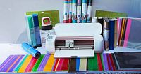 Cricut Maker machine with vinyl sheets laid out in front and rolls and mats behind