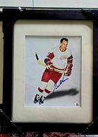 Framed color photograph showing Gordie Howe in Detroit Red Wings hockey uniform, skating with a stick and signed in blue ink.
